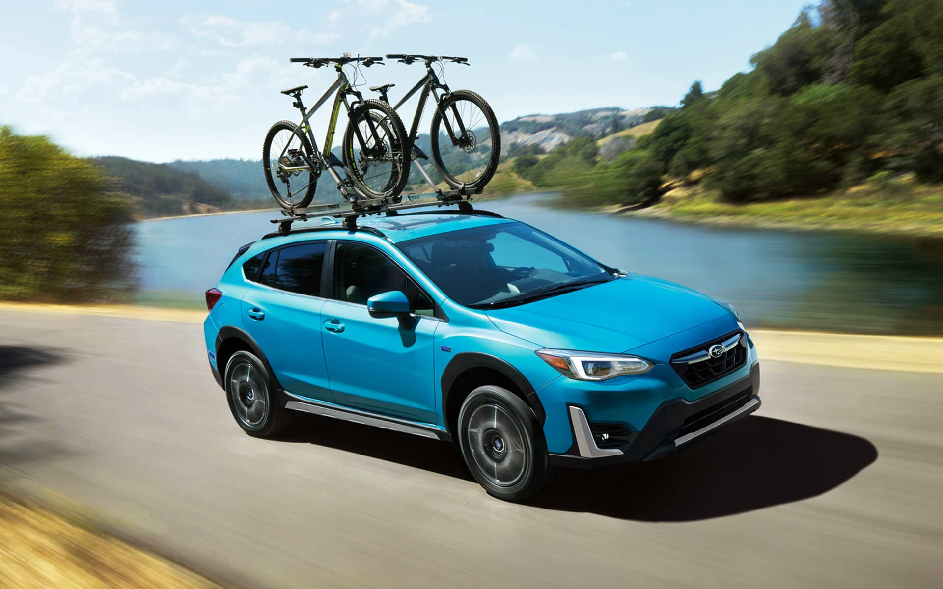 A blue Crosstrek Hybrid with two bicycles on its roof rack driving beside a river | Vann York Subaru in Asheboro NC